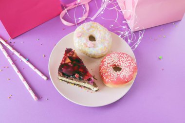 A piece of chocolate cake with cream and berries and sweet donuts on a plate, holiday gifts and illumination, on a pink and purple background