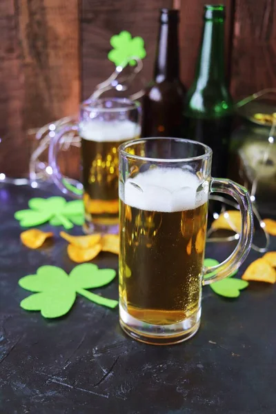 Patrick\'s Day, foamy beer in glass mugs and a bottle, chips and cookies, green shamrocks and gold coins, on a wooden background, party, St. Patrick\'s Day celebration