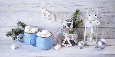 İki fincan sıcak çikolata, marşmelov ve Noel süslemeleri, aydınlatma, beyaz ahşap arka planda.