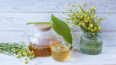 Cam çaydanlıkta papatya çiçekleri olan şifalı bitkisel çay, bir buket taze çiçek, ahşap ışıklı arka planda..