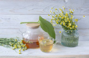 Cam çaydanlıkta papatya çiçekleri olan şifalı bitkisel çay, bir buket taze çiçek, ahşap ışıklı arka planda..