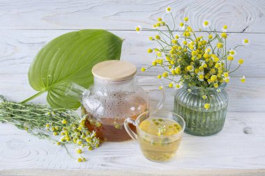 Cam çaydanlıkta papatya çiçekleri olan şifalı bitkisel çay, bir buket taze çiçek, ahşap ışıklı arka planda..