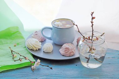 Bir fincan kremalı sıcak çikolata ve mavi ahşap bir masadaki cam vazoda çiçek açan bir kayısı dalı..