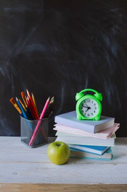 Okula, kırtasiyeye, çalar saate, kitap yığınına karatahta arkaplanlı tahta masaya..