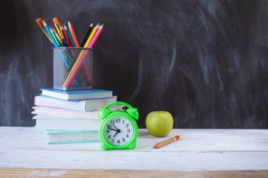 Okula, kırtasiyeye, çalar saate, kitap yığınına karatahta arkaplanlı tahta masaya..