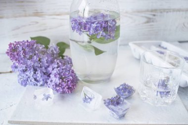  Leylak çiçekleri, buz küpleri ve arka planda bir bardak su ve vazoda leylak çiçekleri.