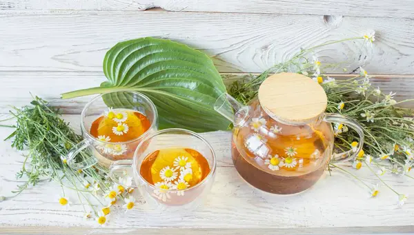 Cam çaydanlıkta papatya çiçekleri olan şifalı bitkisel çay, bir buket taze çiçek, ahşap ışıklı arka planda..