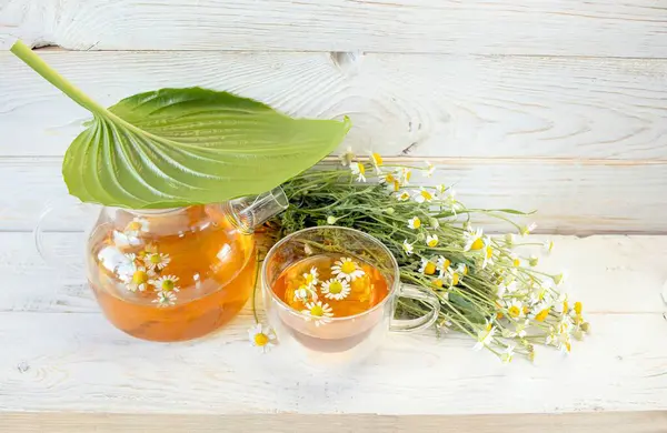 Cam çaydanlıkta papatya çiçekleri olan şifalı bitkisel çay, bir buket taze çiçek, ahşap ışıklı arka planda..