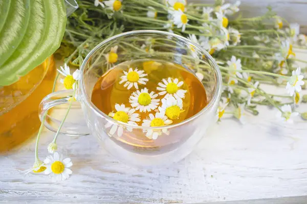 Cam çaydanlıkta papatya çiçekleri olan şifalı bitkisel çay, bir buket taze çiçek, ahşap ışıklı arka planda..