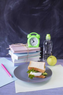 Okul konseptine geri dönelim. Masanın üzerinde okul yemeği, çalar saat ve kırtasiye malzemeleri var. Bir sandviç, bir elma, bir şişe su ve ders kitabı..