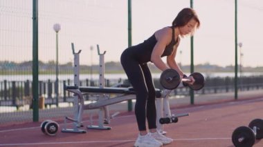 Fit woman strengthens muscles of back lifting heavy barbell standing on red flooring of sports ground at sunrise. Active female person in sportswear does exercise with weight to improve body shape