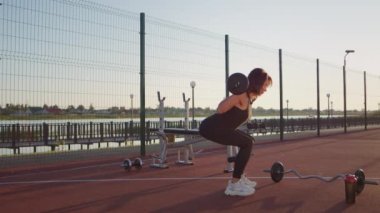 Sportive woman does squats with weight on sports ground on city stadium at sunrise. Motivated mature sportswoman enjoys intense body training with barbell to pump up body muscles in summer morning