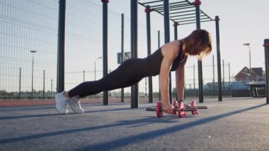 Motivated woman in sportswear does push-ups putting hands on dumbbells on sports ground illuminated by sunrise light in early morning. Sportive lady enjoys effective training to strengthen body