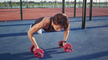 Mature woman pumps up arm muscles doing push-ups with dumbbells on blue flooring of sports ground. Lady in sportswear enjoys taking care of health and soul doing regular exercises in morning