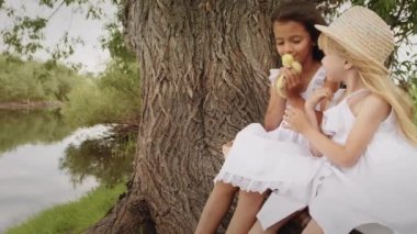 Cute little girls kiss and stroke furry yellow duckling on cloudy summer day in countryside. Brunette and blonde friends sit near big tree growing on river bank against water showing love for bird