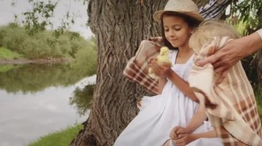 Adorable little girls hold furry yellow duckling in hands sitting near old tree growing on river bank against water. Caring mother covers children with warm plaid on cool summer day in countryside