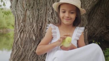 Little girl with smile on face strokes hatched yellow duckling leaning on big tree growing on river bank against water. Caring child holds bird in hands enjoying summer day in countryside closeup