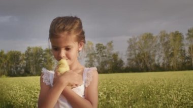 Smiling little girl takes care of furry yellow duckling standing on green field against lush trees under grey cloudy sky. Caring child kisses and strokes bird on summer day in countryside closeup