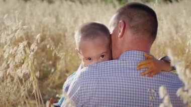 Little boy with Down syndrome hugs father looking in camera near golden spikelets waving in wind. Man expresses love for son with intellectual disability sitting in rural field on summer day closeup