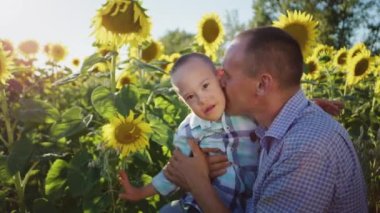 Man kisses little son with Down syndrome sitting among blooming sunflower field illuminated by bright sunlight. Caring father enjoys spending summer vacation with sick child in countryside closeup