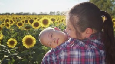 Caring mother kisses cheek of little son with Down syndrome expressing love. Sweet boy rests in woman arms enjoying walk across blooming sunflower field in countryside on sunny summer day closeup