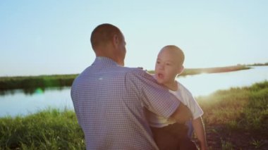 Caring father holds little son with Down syndrome on lap sitting on river bank contemplating bright sunset. Smiling man and disabled boy with mental disability spend time in countryside in summer