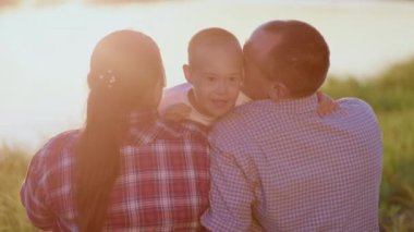Loving parents hold little disabled son sitting on river bank illuminated by sunset light. Happy preschooler boy with Down syndrome smiles hugging mother and father on summer walk closeup