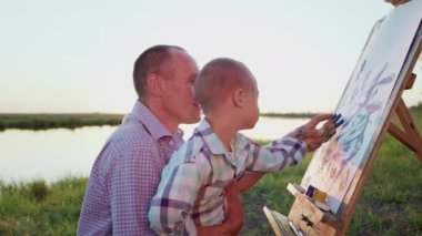Smiling father enjoys teaching little son with Down syndrome to paint on easel canvas with fingers sitting on grassy river bank. Caring man spends time with curious disabled preschooler boy closeup