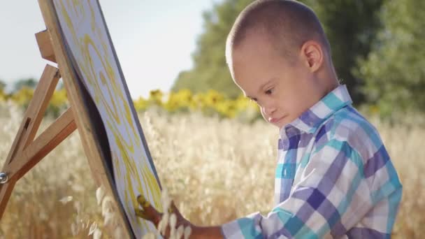 下症候群の子供は 背景のぼやけた金色のフィールドに立っているキャンバスのイーゼルに指で黄色の太陽を描きます 小さな男の子は田舎の閉鎖で晴れた夏の日に芸術と農村の自然を楽しんでいます — ストック動画
