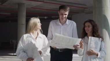 Successful civil engineer talks to female investors walking at commercial building construction site with concrete beams. Man and women discuss project plan looking at paper drawings of skyscraper