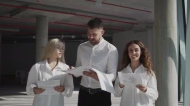 Professional civil engineer inspects industrial construction site of high-rise building with female colleagues. Young specialists check condition of area looking at paper drawings and project plan