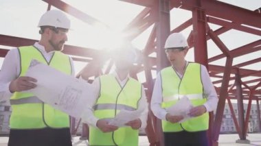 Team of three smiling builder wearing safety uniform discuss construction blueprint walking along industrial facility outdoors in sun light. New project development, real estate engineering works