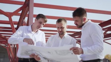 Group of successful investors standing at commercial building construction site against structural metal beams under blue sky. Young male colleagues discuss new project studying big paper drawing