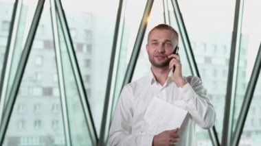 Professional civil engineer talks to client on smartphone against bright panoramic window of skyscraper. Young bearded man discusses working details standing at industrial construction site closeup