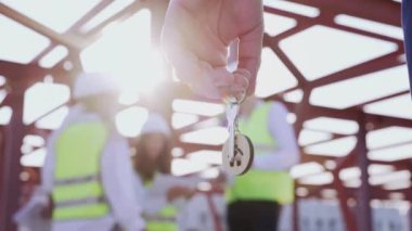 Male investor holds key to apartment against group of professional engineers standing at commercial construction site with metal beams. Purchase of property in high-rise building object closeup