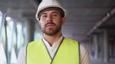 Bearded man walks at construction site of high-rise building near panoramic window. Portrait of young civil engineer looking in camera and turning head aside on blurred background close view