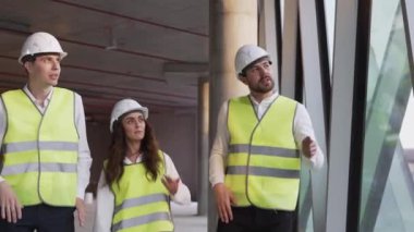 Three smiling professional engineer in safety wear discuss future real estate construction and new project plan, stand on building facility indoors. Architectural works development by contractor team