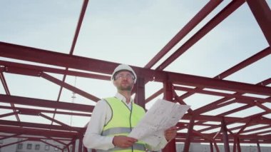 Professional civil engineer in hardhat holds paper drawing inspecting area. Young bearded man stands at industrial construction site of high-rise building against structural metal beams low angle shot