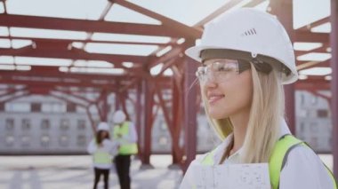 Portrait of young female civil engineer in goggles turning to camera smiling against busy colleagues. Professional specialist stands at construction site with formwork frames on sunny day closeup