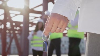 Hand of female client holding key to new apartment at modern building object on blurred background. Woman and professional engineers stand at industrial construction site with metal beams closeup