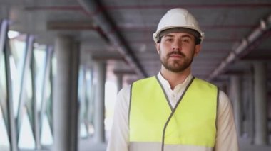 Portrait of successful young architect in hardhat and safety vest looking in camera on blurred background. Bearded male specialist walks on construction site of building with panoramic windows closeup