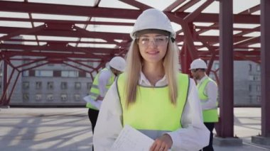 Cheerful woman in protective helmet smiles looking in camera standing at construction site of high-rise building. Portrait of successful female contractor against colleagues working on project closeup