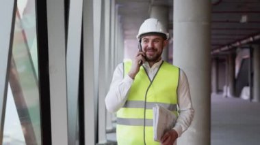 Happy smiling male engineer architect walking along building facility, talking real estate works plan with team using mobile phone indoors. New project development together for successful future deal