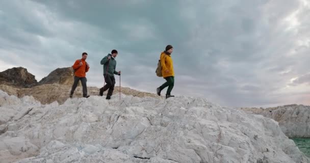 3人の若いハイカーが岩山に沿って湖水に行き 崖の上に立っている トレッキングウォーク 観光の発見 野生の自然での旅行の成功 — ストック動画