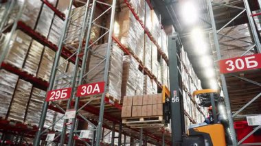 A worker on a forklift lifts boxes of goods and places them on a shelf of a rack in a large modern distribution center. Warehouse filled with cardboard boxes on shelves A shipping and supply chain