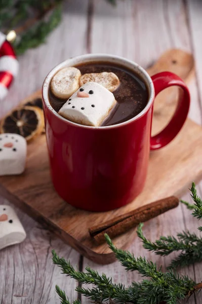 Red mug with hot chocolate and melted marshmallow snowman, rustic wooden festive background.