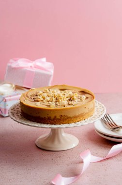 Close up caramel cheesecake on cake stand with gift boxes and pink background.