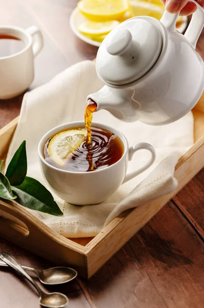 Black tea, pouring tea from tea pot in a white, ceramic cup. Wooden, rustic background.