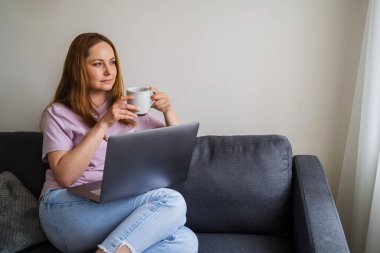 İnternette çalışan, kanepede oturan, oturma odasında kahve içen ve dizüstü bilgisayar kullanan bir kadın..