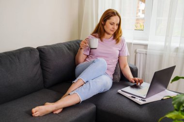 İnternette çalışan bir kadın, oturma odasında bir koltukta oturuyor ve dizüstü bilgisayar kullanıyor. 